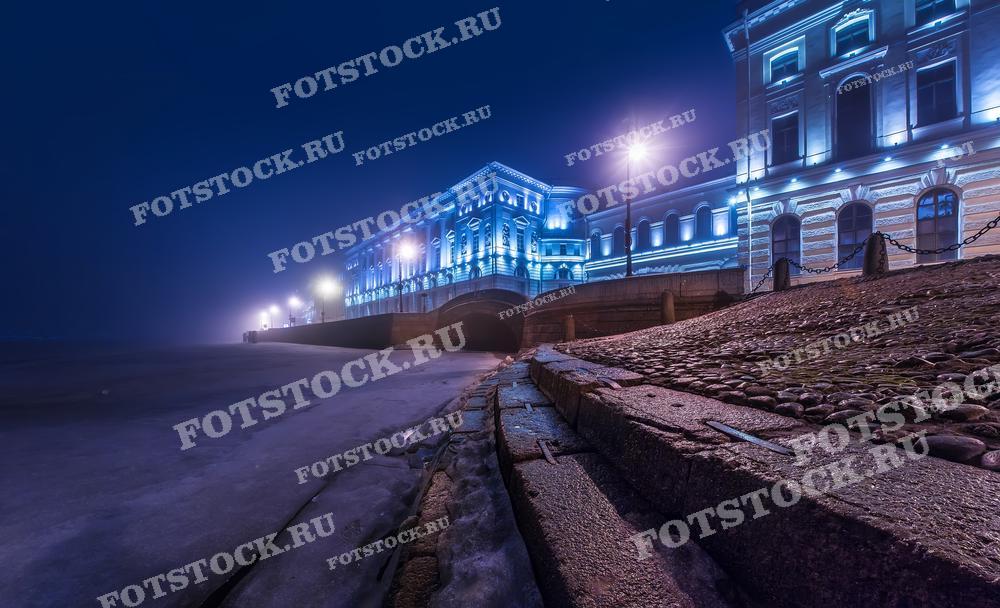 На фотографии изображена набережная Санкт-Петербурга ночью зимой. Видны освещенные здания и замерзшая река. Фото может использоваться для туристических буклетов, открыток и рекламных материалов.